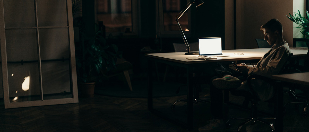 Auf dem Bild sieht man einen Mann in einem dunklen Zimmer an einem Schreibtisch sitzen. Das einzige Licht ist eine Schreibtischlampe.