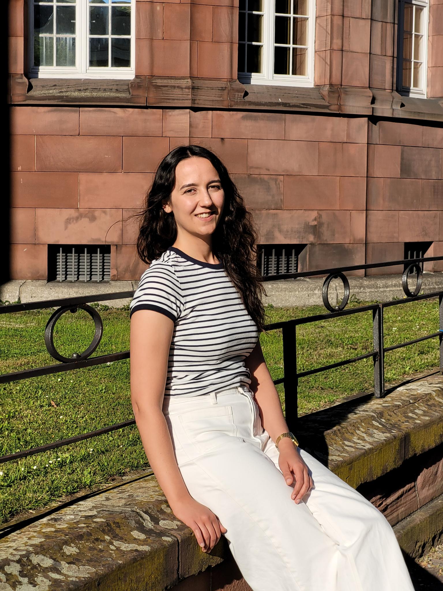 signal-2025-05-10-20-59-15-565 Auf dem Bild ist eine junge Frau in heller Kleidung zu sehen, die vor dem Universitätsgebäude KG I auf einer Mauer sitzt.