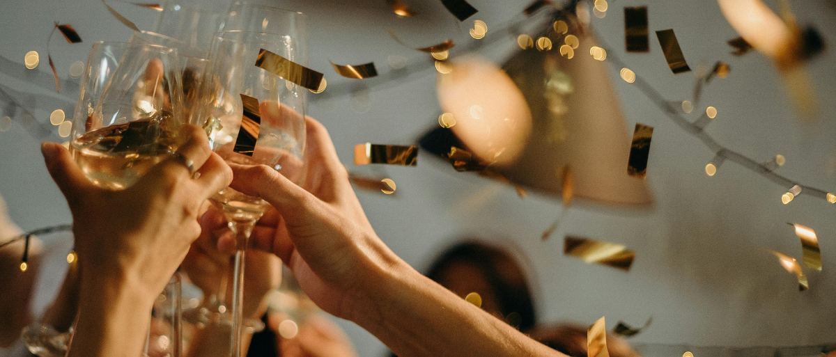 In diesem Bild stoßen mehrere Hände mit Sektgläsern an und es fliegt goldenes Konfetti durch die Luft.