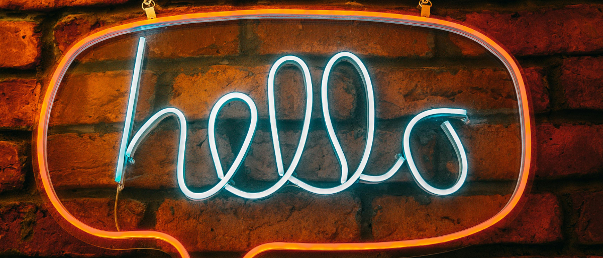 Man sieht einen Leuchtschriftzug "hello" in hellblau in einer Orangengen Sprechblase. Im Hintergrund ist eine rote Backsteinmauer zu sehen.