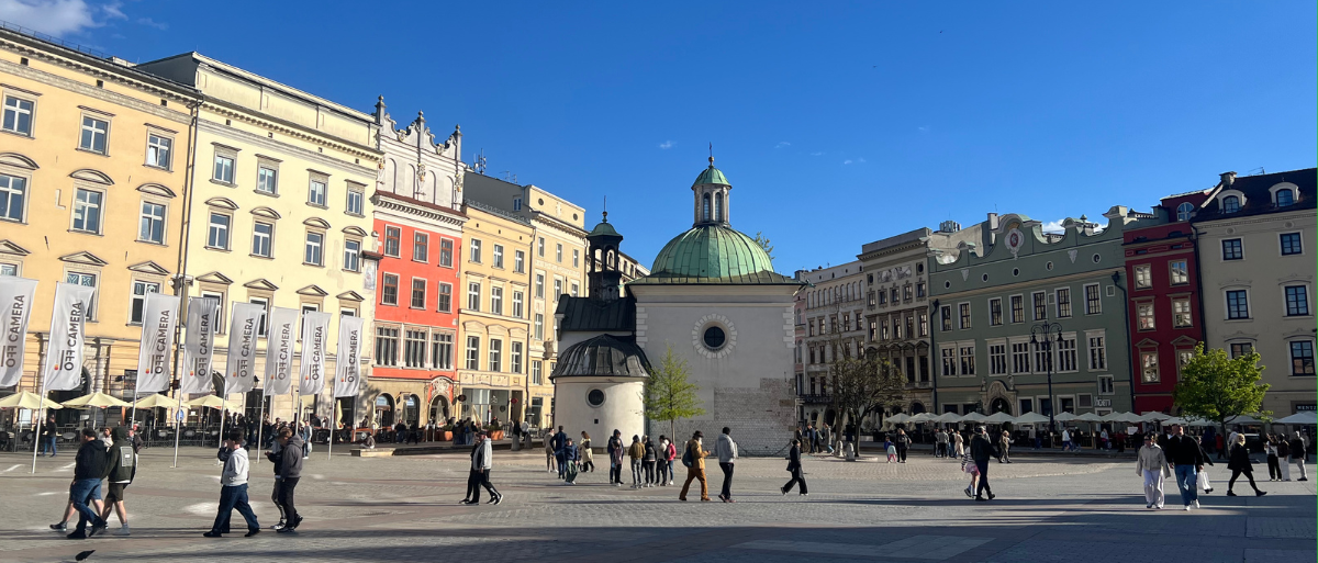 Ein Blick auf eine Plaza in Krakau, man sieht Menschen, die über einen großen Platz laufen und bunte, schmucke Hausfassaden im Hintergrund.