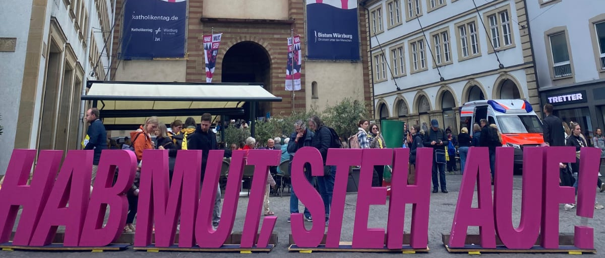 Auf dem Bild steht in pinken Lettern: Hab Mut, steh auf! Im Hintergrund ist der Würzburger Dom zu sehen.