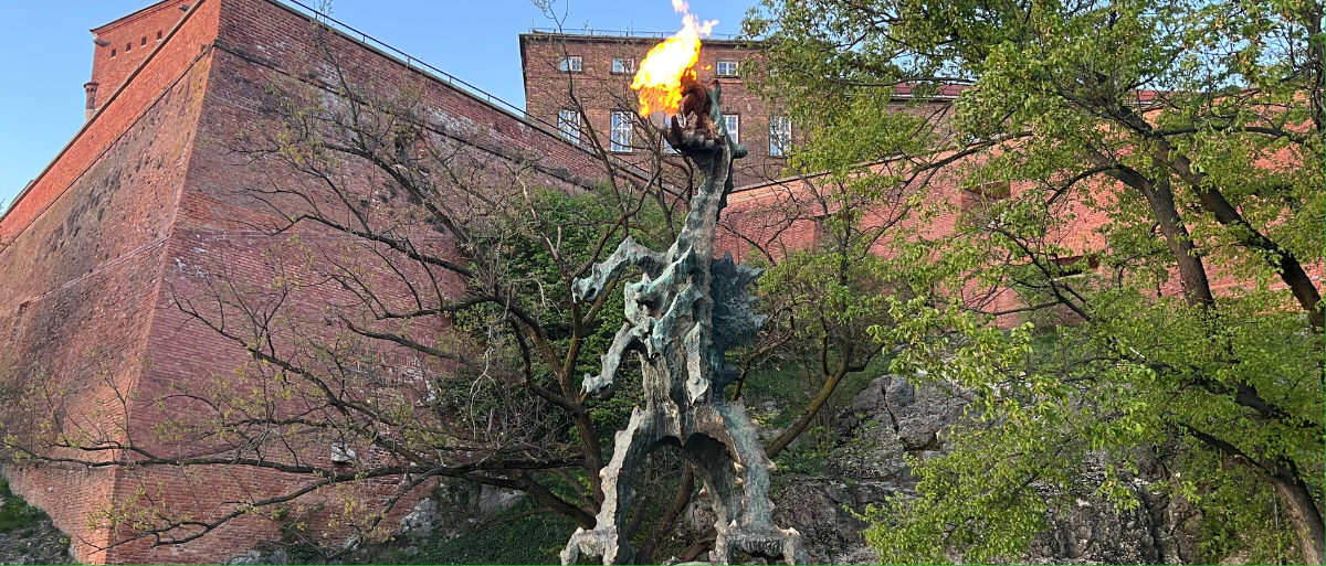 Man sieht eine feuerspuckende Statue eines Drachen (echtes Feuer). Die Statue steht auf einem großen Stein. Im Hintergrund ist die Wand eines Backsteingebäudes zu sehen, außerdem gibt es grüne Bäume im Hintergrund.