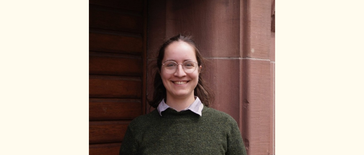 100 Jahre Caritaswissenschaft (1) Auf dem Bild ist Lisa Baumeister zu sehen, wie sie vor dem roten Universitätsgebäude der Uni Freiburg in die Kamera lächelt.